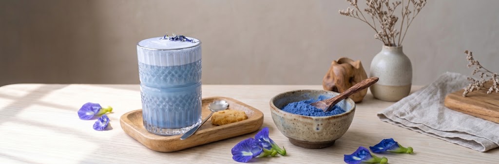 Kittelbloempoeder in een kom naast een glas blauwe matcha, met een vaas en kittelbloem eromheen